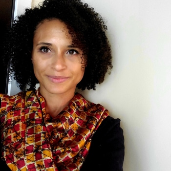 Jessica Horvath Williams is a light-brown-skinned black woman with dark curly hair. She is wearing a black shirt with a multi-colored red and orange scarf, and is standing in front of a white wall.