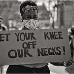 Black woman at a protest in a facemask holding a sign Get Your Knee Off Our Necks