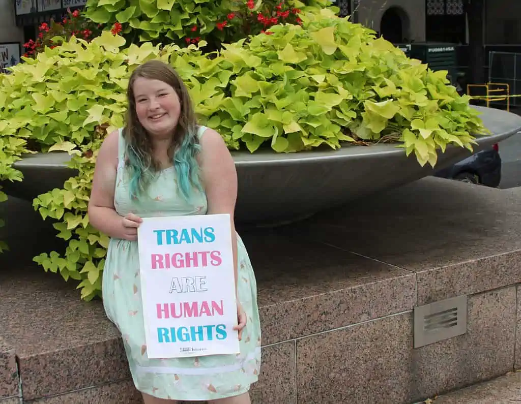 A young white woman with medium-length brown hair that fades into teal sits in front of a large plant. She is wearing a knee-length light green sundress with ice cream cones on it. She is holding a sign reading 'trans rights are human rights' in alternating colors of the trans pride flag.