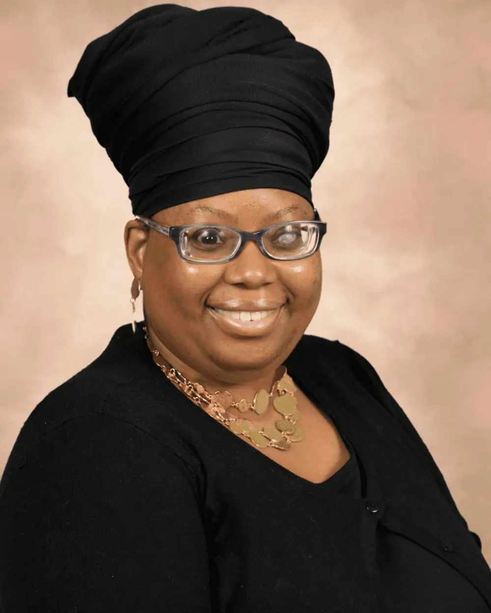 Carla, a Black woman with a tall black headwrap, glasses, and gold jewelry, smiles at the camera.