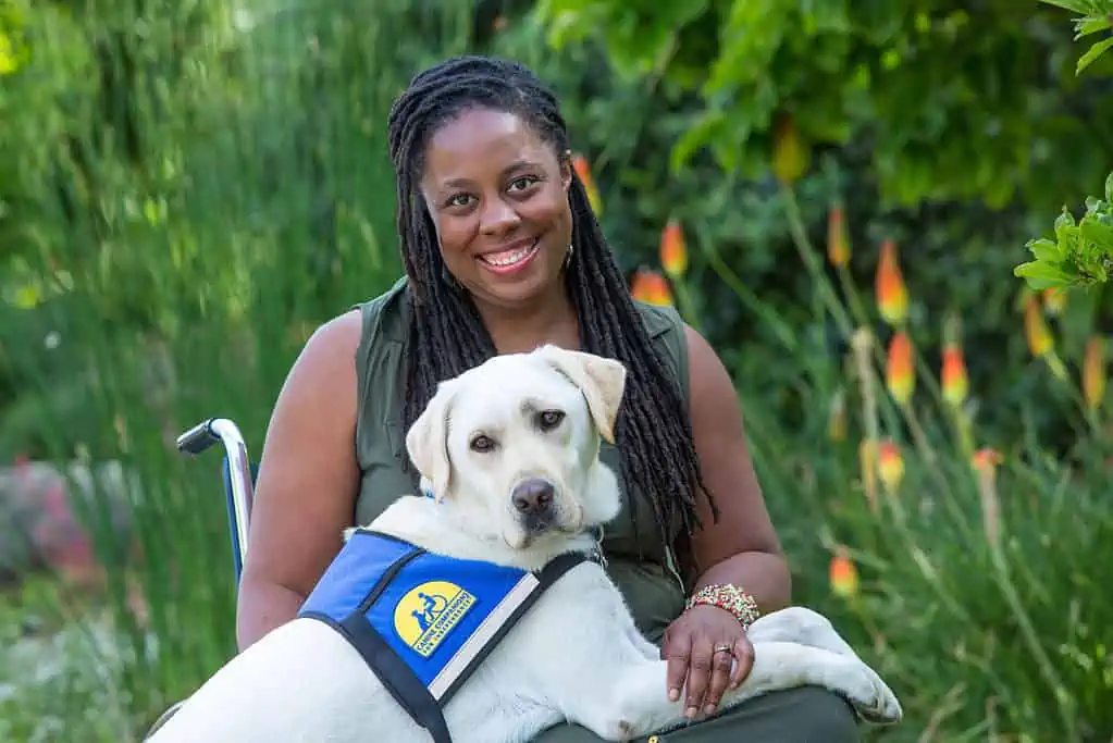 India Harville, African American female with long black locs, seated in her manual wheelchair wearing a long sleeveless green dress.  Her service dog, Nico, a blond Labrador Retriever, has his front paws on her lap.  He is wearing a blue and yellow service dog vest. They are outside with greenery behind them.