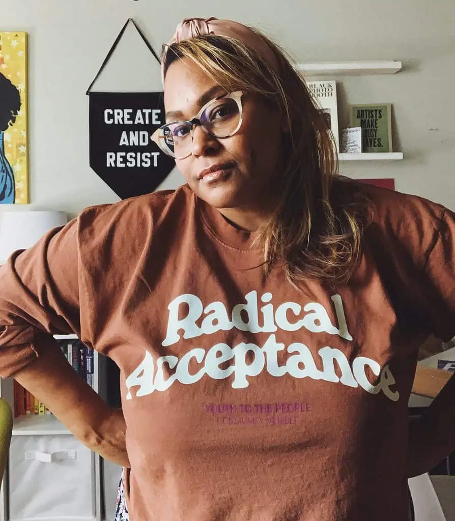 Jen, an Afro Latina Woman with caramel brown skin, staring pensively at the camera. She is standing in a white room, with artwork on the walls. She is wearing cream colored cat eye glasses, her brown/blonde hair falls on her shoulders, she is wearing a pink headband and a copper colored t-shirt that says radical acceptance in bold white lettering. 