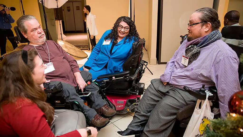Rahnee Patrick (third from left, in the blue jacket), a Thai-white woman in a power wheelchair, smiling and speaking to three other people using wheelchairs.