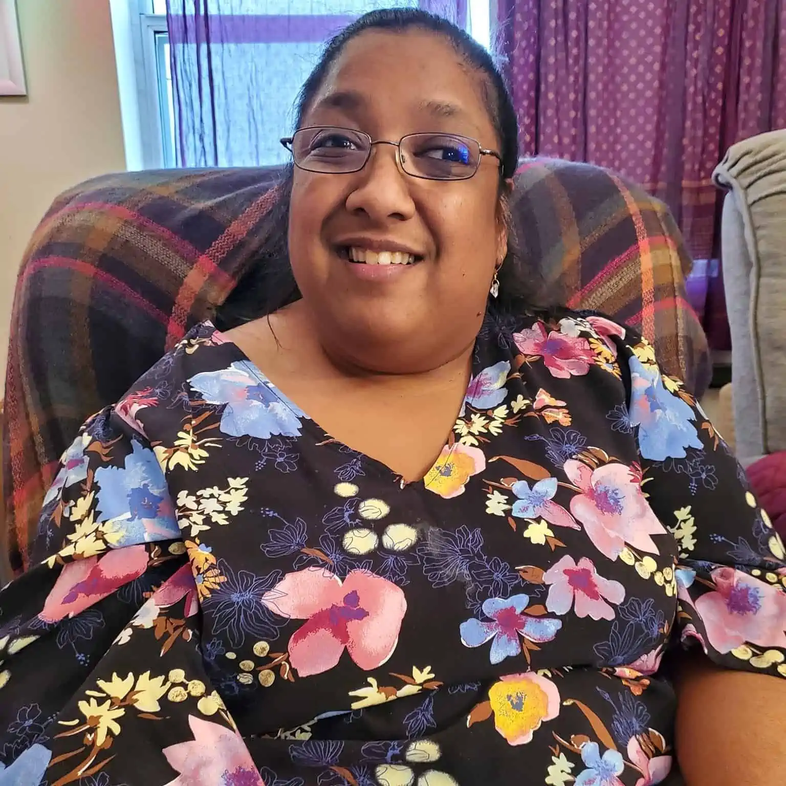Sarah is a dark-skinned woman sitting in her living room recliner. Visible on the back of the recliner is a gray shawl with a bright plaid pattern. A window is behind her, with long dark purple curtains to the right. She is wearing black wire-rimmed glasses that have a purple tint. Sarah also is wearing small silver earrings that shimmer as well as a shirt that has a floral pattern over a navy blue background. The flowers are light blue, purple and yellow. She’s smiling widely, and her eyes are actually open, which is one reason she chose this photo.