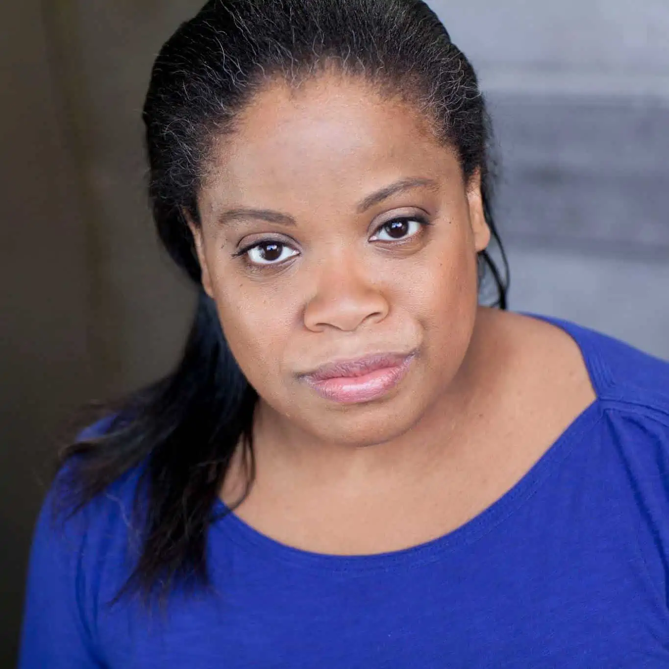 Terri - A brown skinned Black woman with shoulder length straight dark hair, greying around the edges and swept over one shoulder, looks straight into the camera with a mostly serious expression and slight smile. She is wearing an indigo shirt.