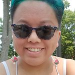 Nancy Yang, mad queer HMoob womxn, wearing short teal hair and black sunglasses and white tanktop.