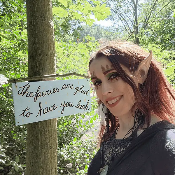 Kayley Whalen autistic woman wearing elf ears and elven face paint in a black pagan dress standing in front of a sign reading "the faeries are glad to have you back" in the forest