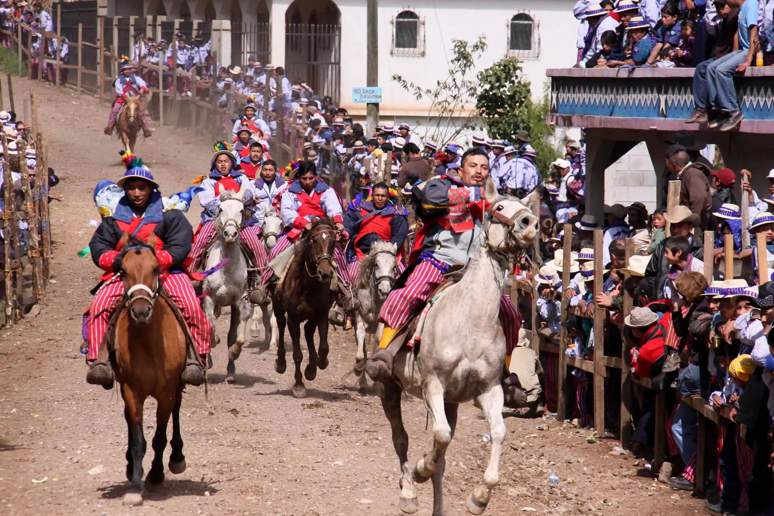 Horse racing with men in traditional Mam Maya clothes riding horses down a narrow track thronged with people.