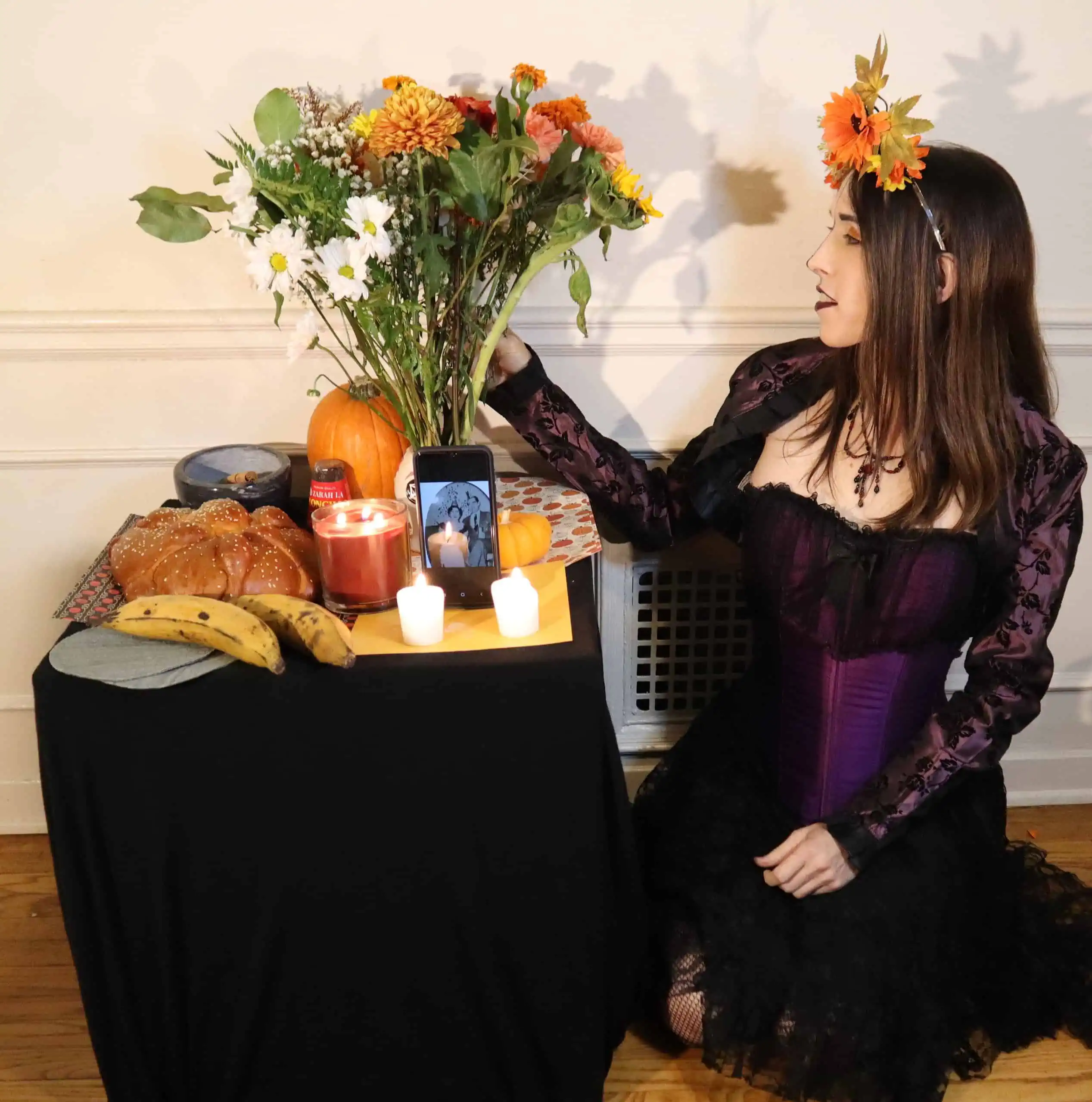 An altar (ofrenda) with flowers, candles, food, and plantains. Kayley Whalen, a Latina trans woman, is wearing a corset and flowers in her hair and kneeling at the altar.