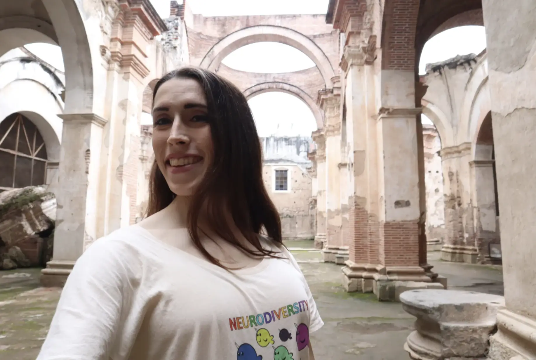arched ruins of Spanish Catholic church in original colonial capital of Guatemala in Antigua. Smiling Latina woman Kayley Whalen is wearing a "Neurodiversity is for Everyone" t-shirt with the flappy narwhals created by the Ed Wiley Autism Acceptance Lending Library.