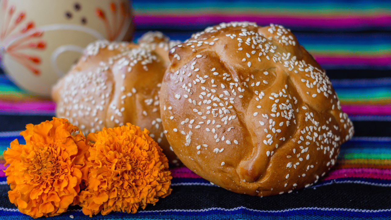 A large loaf of sweet bread with bone-like shapes on top and sesame seeds, next to orange marigolds. It is on top of a bright textile pattern.