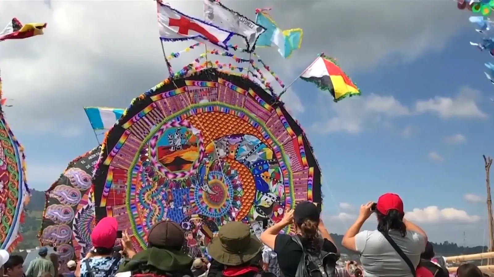 Giant bright multicolor circular/octogonal woven kite with neon fringed flags flapping in the wind. One flag is the Guatemalan flag. Other giant kites are visible near it.