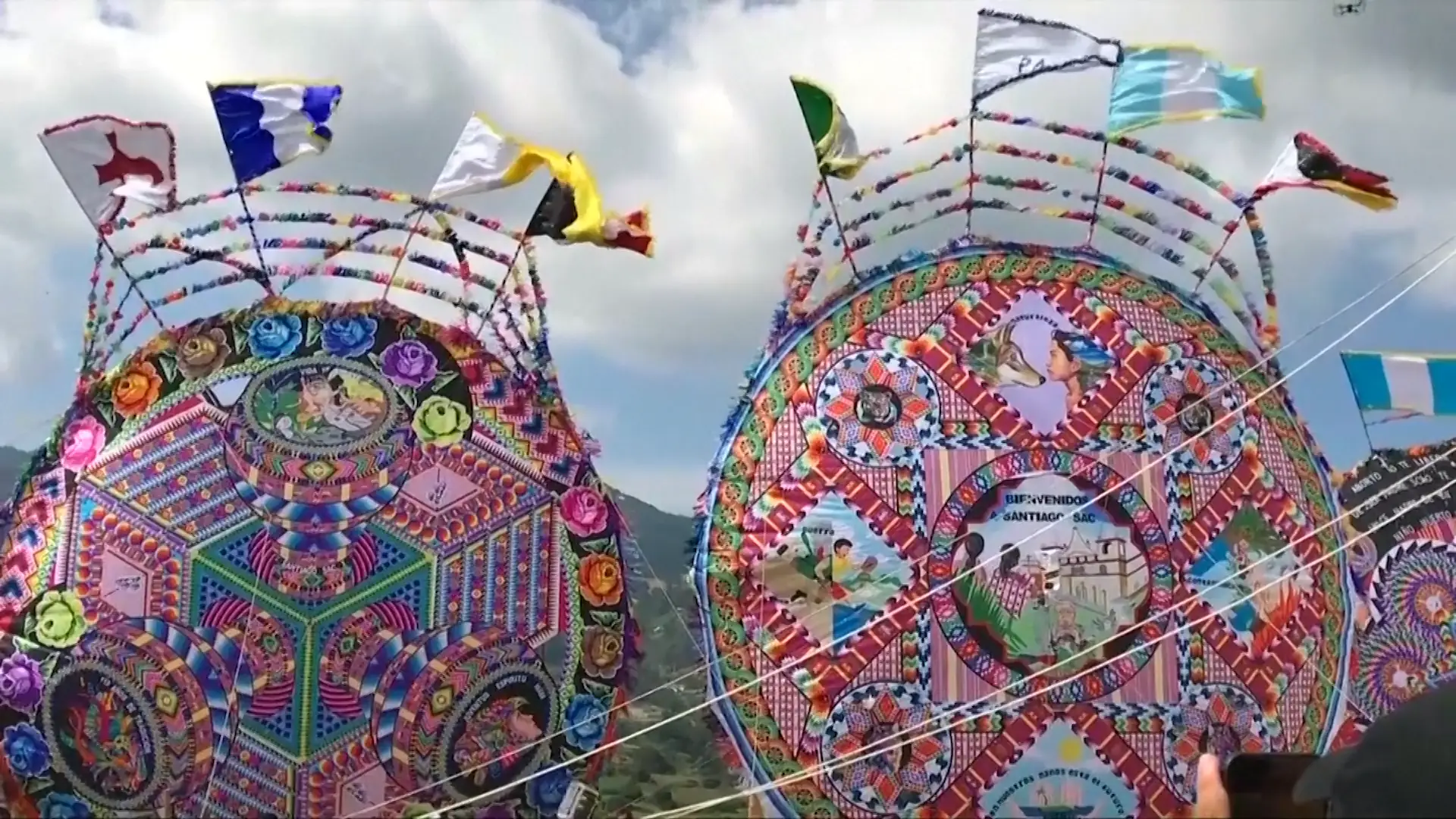 Two-story tall kites anchored to the ground in Santiago Guatemala. The kites are made with traditional multicolor Maya textile patterns and art depicting life in Guatemala. The kites are surrounded by a fringe of fabric and various flags blowing in the wind.