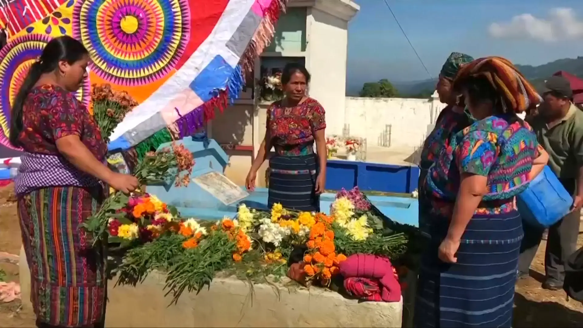 Maya women in traditional clothing placing orange marigolds and other flowers on a raised grave.  A giant kite is visible over the grave, as are additional graves.