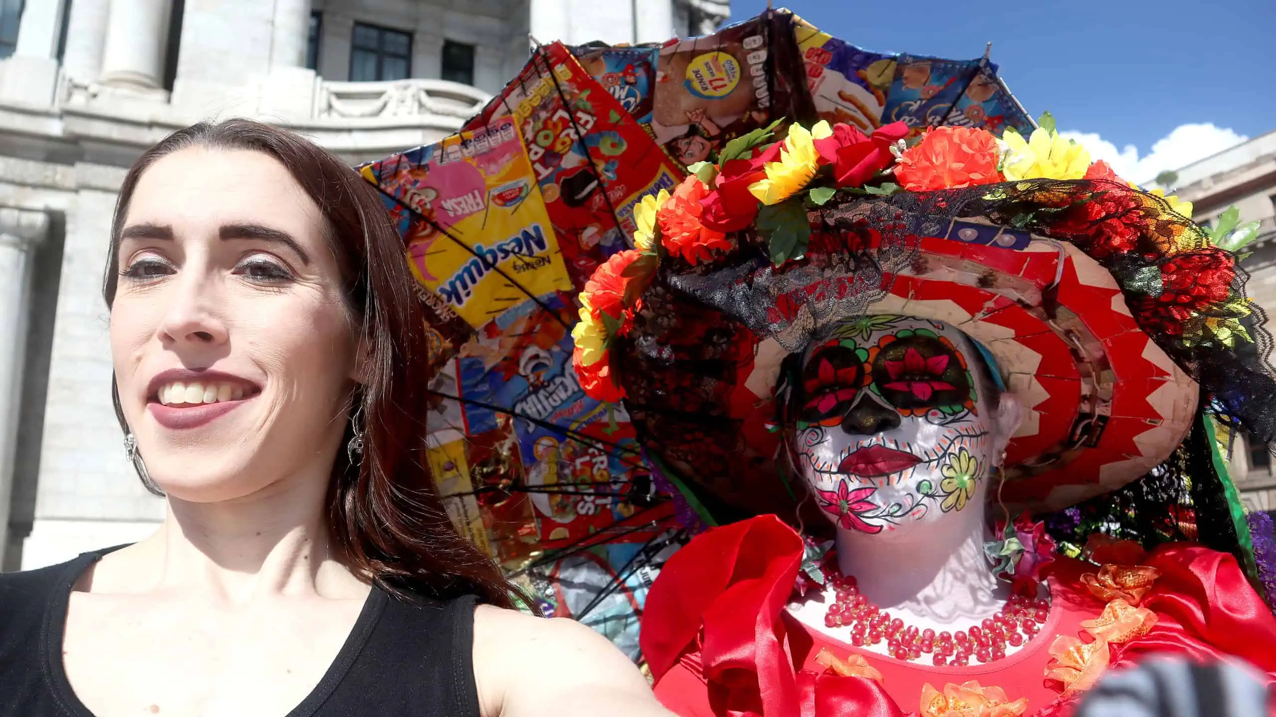 A Mexican woman in skull face paint with a fancy hat and parasol made from junk food cereal boxes with US brands poses for a selfie with Kayley Whalen.