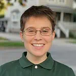 smiling white nonbinary person with short hair and thin glasses wearing a teal button-up