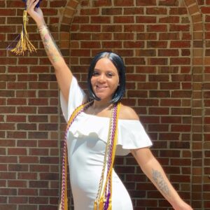 Pictured is Kyara in front of a brick wall. She is smiling, wearing a white dress with graduation cords around her neck. Her right arm is holding up a graduation cap and tassel.