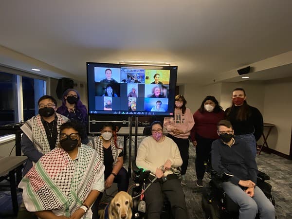 Lydia XZ Brown, Sharon daVanport, Kayla Rodriguez, and other participants at US Gender and Disability Justice Alliance convening gathered in conference room and in a Zoom meeting. Everyone is wearing masks.