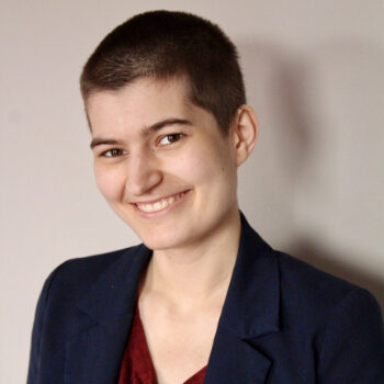 An image of an androgynous Italian person smiling at the camera. Their hair is a dark brown buzz cut, and they are wearing a navy blazer over top of a dark red shirt.