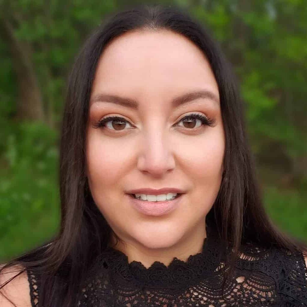 A close-up photo of Jules, a light skinned Indigenous woman with brown hair, brown eyes, neutral makeup, and a light smile. She is outside with greenery in the background, wearing a black high-necked lace dress.