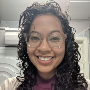 Rieko is facing forward, smiling into the camera. She has brown skin, curly hair, and is wearing pink glasses and a beige cardigan over a burgundy turtleneck.